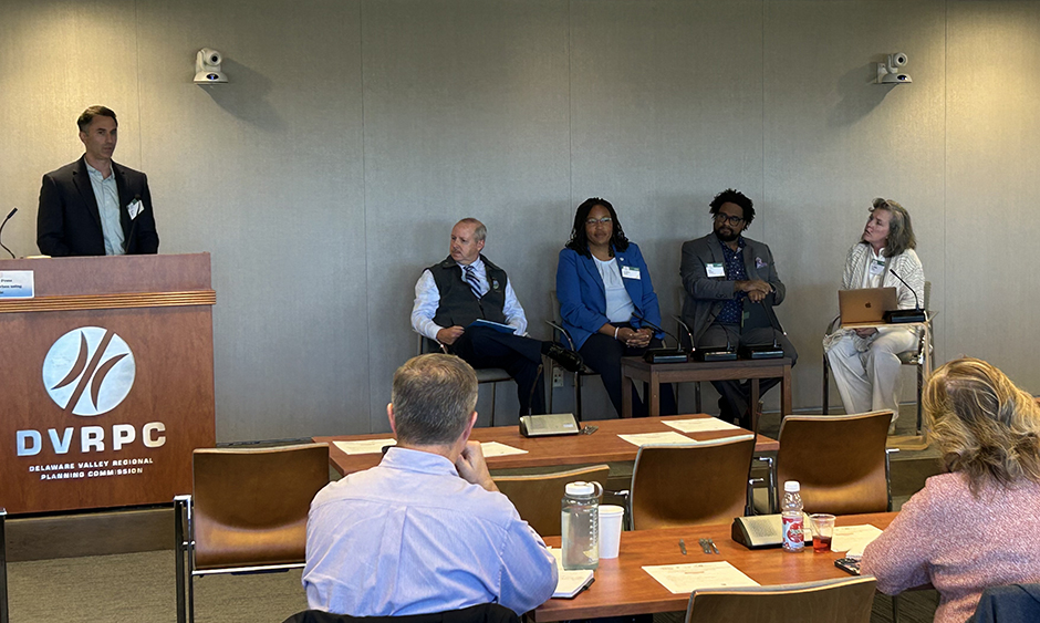 A person speaks at a podium. To their right is a group of four panelists