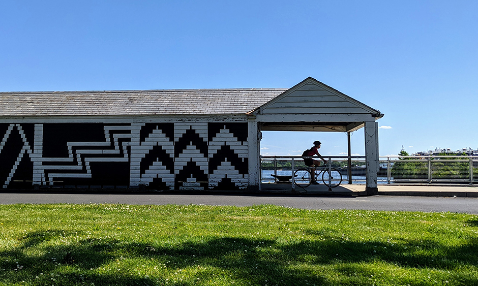 A bicyclist riding on a path in the Philadelphia Navy Yard is framed by a gazebo at the end of a building covered in murals