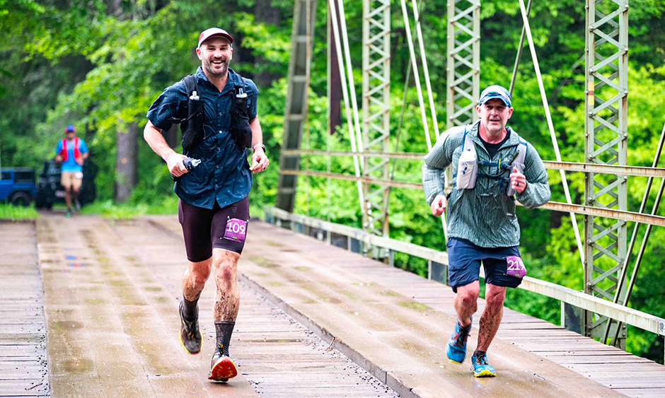 Two people running over a bridge in a wooded area