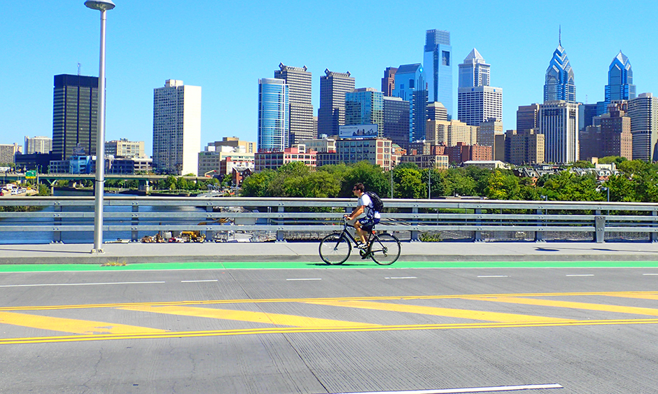 A person rides their bike on a bridge over the Schuylkill River in Philadelphia. The city skyline is in the background.